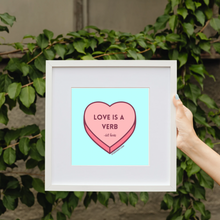 Load image into Gallery viewer, Framed art print that features a pink conversation heart with the words “LOVE IS A VERB - bell hooks” centered on it in a cranberry font on a complementary light blue background.
