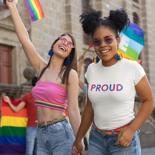 Two femme people walk hand in hand at a Pride event. One is wearing a “PROUD” bisexual PTSFeminist t-shirt and smiling at the camera, the other is smiling while holding a flag in the air.